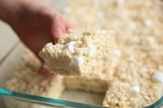 A glass 9x13 dish of cut Rice Krispie Treats with one piece held above by a hand.