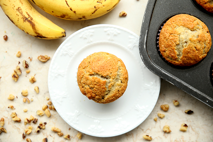 A single muffin on a small plate surrounded by chopped walnuts with two ripe bananas above the plate and the corner of a muffin pan with a muffin in it.