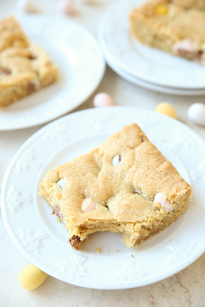 A slice of Easter egg blondies with a bite taken out of the corner sitting on a small white plate. In the background are two more servings of blondies on white plates. Cadbury Mini Eggs are scattered around the plates.