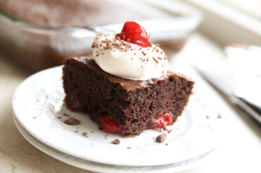 Two white plates stacked on each other with a slice of Easy Black Forest Cake on top topped with a dollop of whip cream, chocolate shavings and one cherry. A 9x13 glass pan of chocolate cake faded in the background.