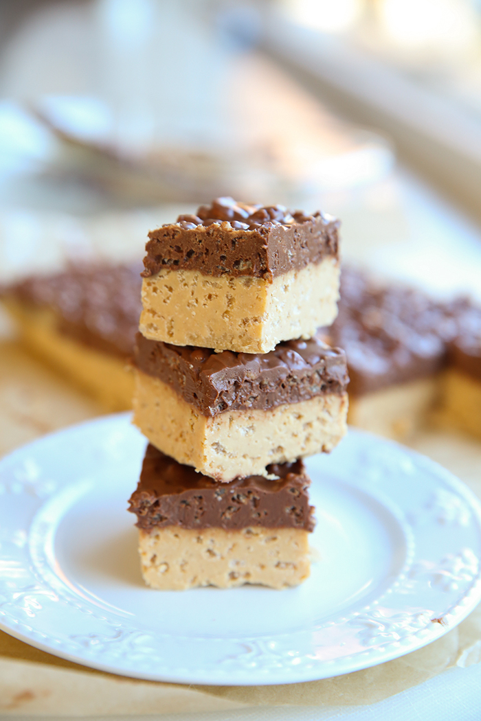 Three slices of peanut butter crunch bars stacked on top of each other on a small white plate with more crunch bars faded in the background.