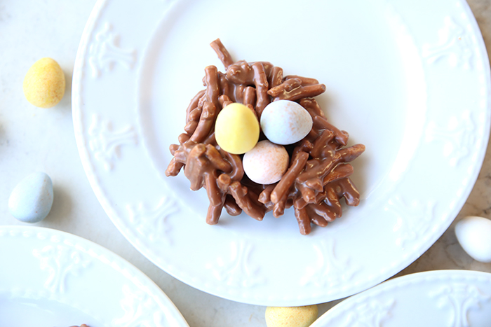 A Birds Nest Cookie sitting on a small white plate with chocolate coated mini eggs surrounding the plate. In the bottom corners are partial pictures of two small plates.