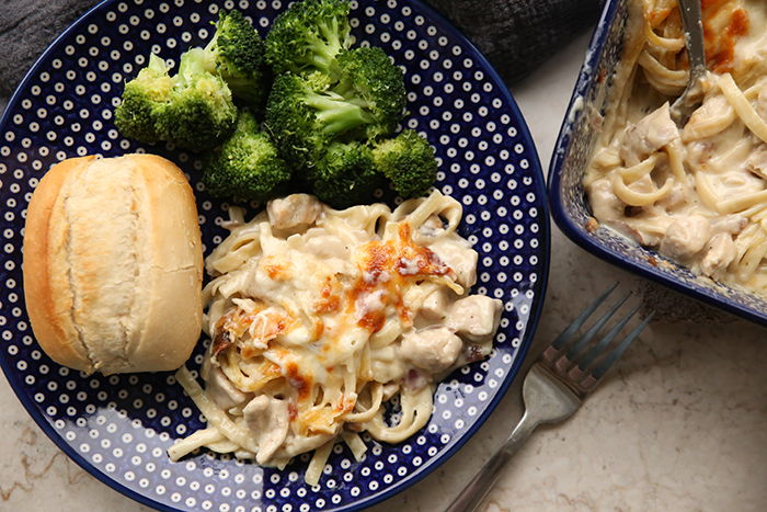 A roll with a serving of broccoli and chicken tetrazzini on a dark blue Polish pottery plate with white dots. In the upper right corner is the corner of a 9x13 Polish potter dish of chicken tetrazzini casserole.