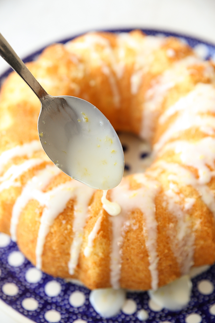 A spoon held above a Whole Lemon Bundt Cake drizzling lemon glaze on the cake which is sitting on a blue and white polka dotted Polish potter plate.