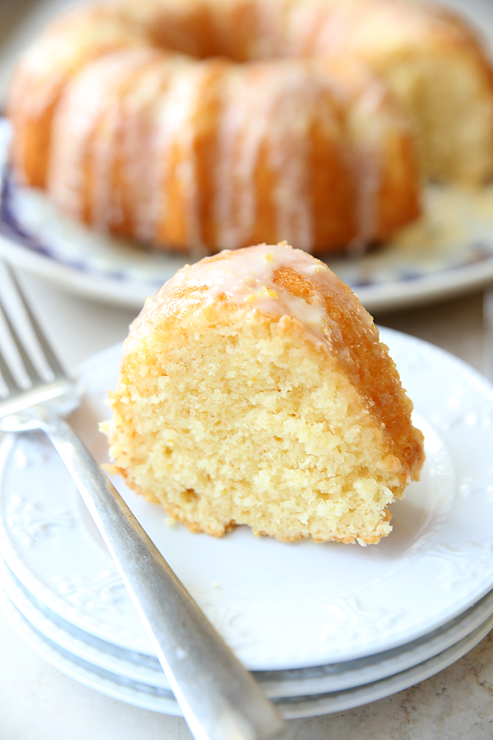 A slice of lemon cake sitting on top of three small plates with a fork lying next to the piece of cake. In the background is a lemon bundt cake with a piece cut out.