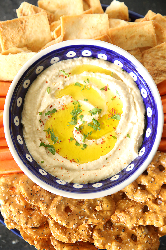 Easy Hummus Recipe in a blue and white peacock patterned Polish pottery bowl surrounded by pretzel and pita chips and sliced carrots.