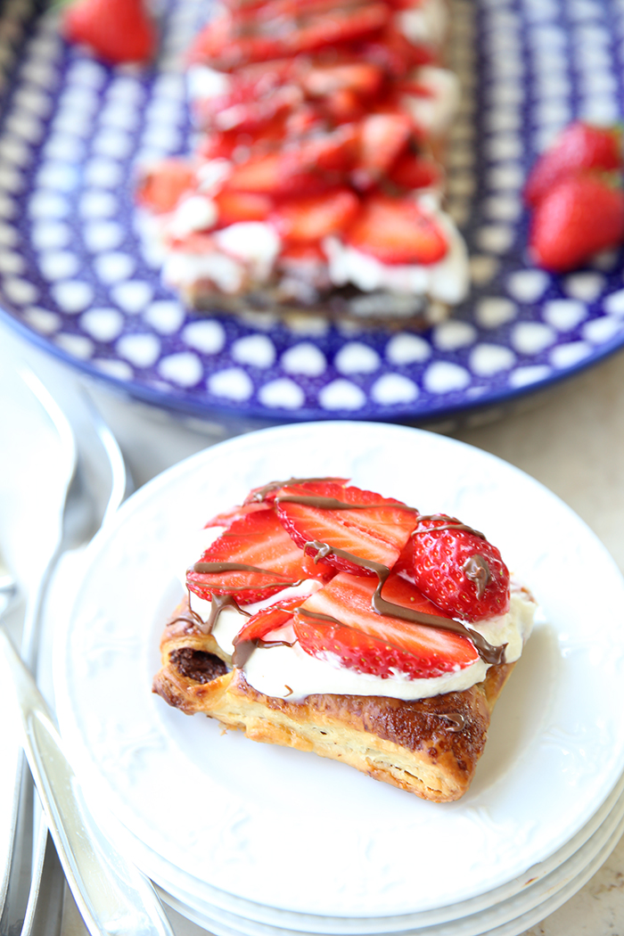 A slice of Strawberry Nutella Puff Pastry Dessert on top of several small plates. Forks are stacked on the left side of the plates. In the upper background is an oval blue white heart patterned dish with the remainder of the dessert with a few strawberries around the dessert.