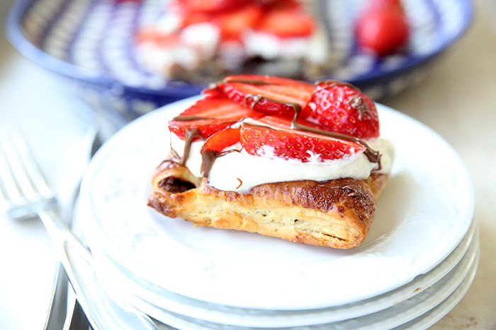 A piece of Strawberry Nutella Puff Pastry sitting on top of four small white plates with a couple of forks on the left. Faded in the back is the puff pastry dessert on a dark blue and heart patterned oval plate.