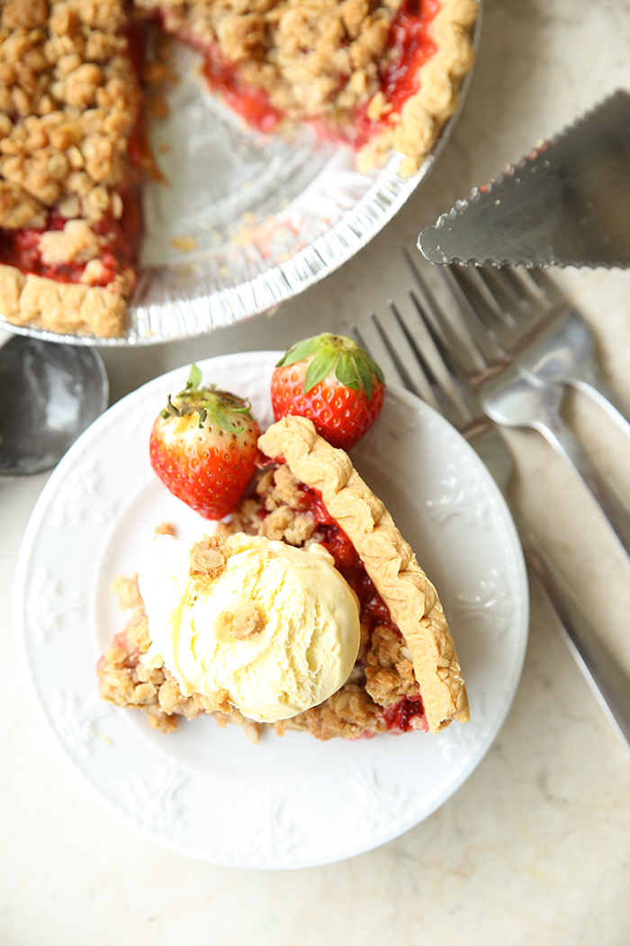 A slice of rhubarb strawberry pie topped with vanilla ice cream sitting on a dessert plate. Next to the plate on the right are three forks and a pie server. Above the pie slice is a strawberry rhubarb pie with a piece cut out.