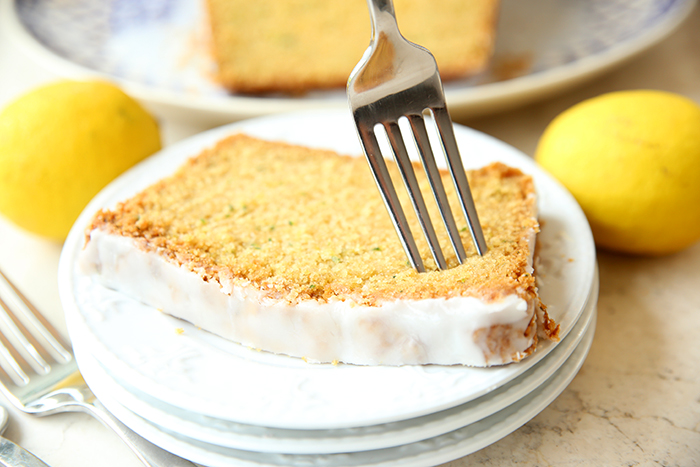 A slice of lemon zucchini bread on three white plates with a fork inserted vertically into it. Two lemons are at the top and right of the plates with a fork head in the bottom left corner. Faded in the upper background is a partial picture of a plate and glazed lemon zucchini bread.