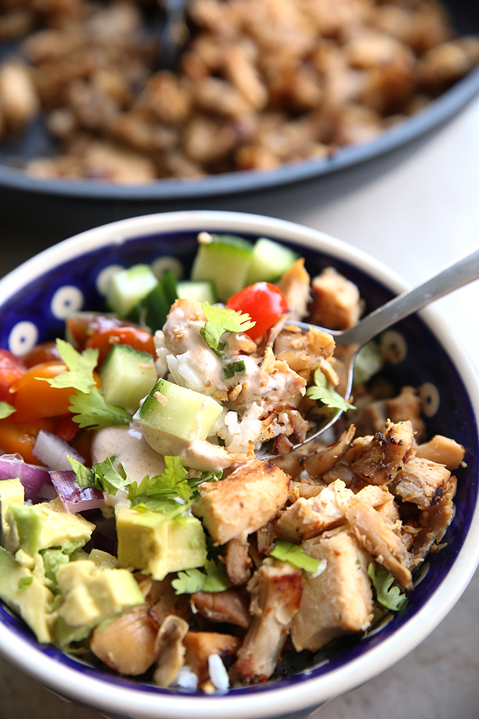 A honey lime chicken bowl with a spoon held above filled with the ingredients of the bowl. Faded in the upper background is a skillet of chopped chicken. 