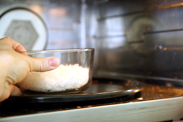 An open microwave with a hand holding a glass bowl of coconut and placing it in the microwave.
