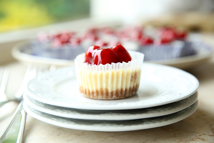 A mini cherry cheesecake on top of three small plates with two forks on the left of the plate. Faded in the upper background are more little cheesecakes on a large plate.