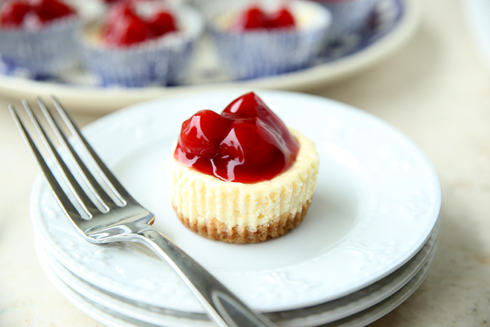 One mini cheesecake and a fork sitting on top of three small plates. In the faded background is a plate of mini cherry cheesecakes.