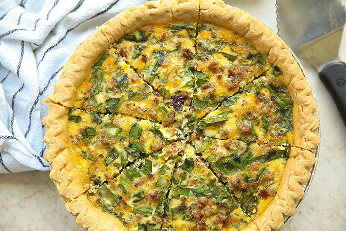 A Sausage and Spinach Quiche cut into 8 pieces with a serving spatula in the upper right corner and a striped tea towel in the upper left corner.
