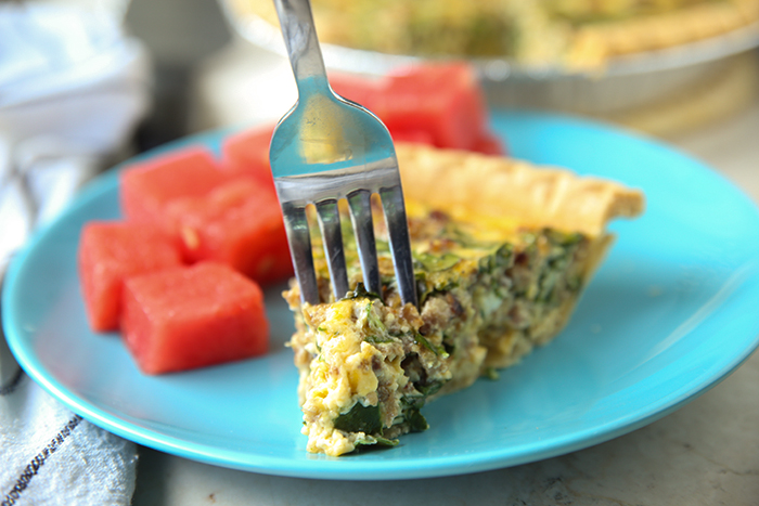 A slice of sausage and spinach quiche with a fork inserted at the tip and several cut up pieces of watermelon all sitting on a blue dinner plate.
