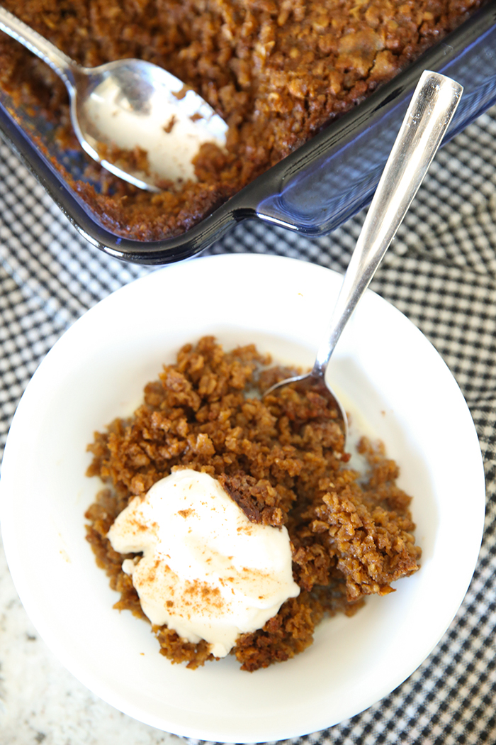 A serving of Gingerbread Baked Oatmeal in a bowl topped with whipped cream and a spoon inserted into the bowl. In the Upper background is a corner of a glass 8x8 dish of baked oatmeal with a spoon in the corner with a serving taken out.
