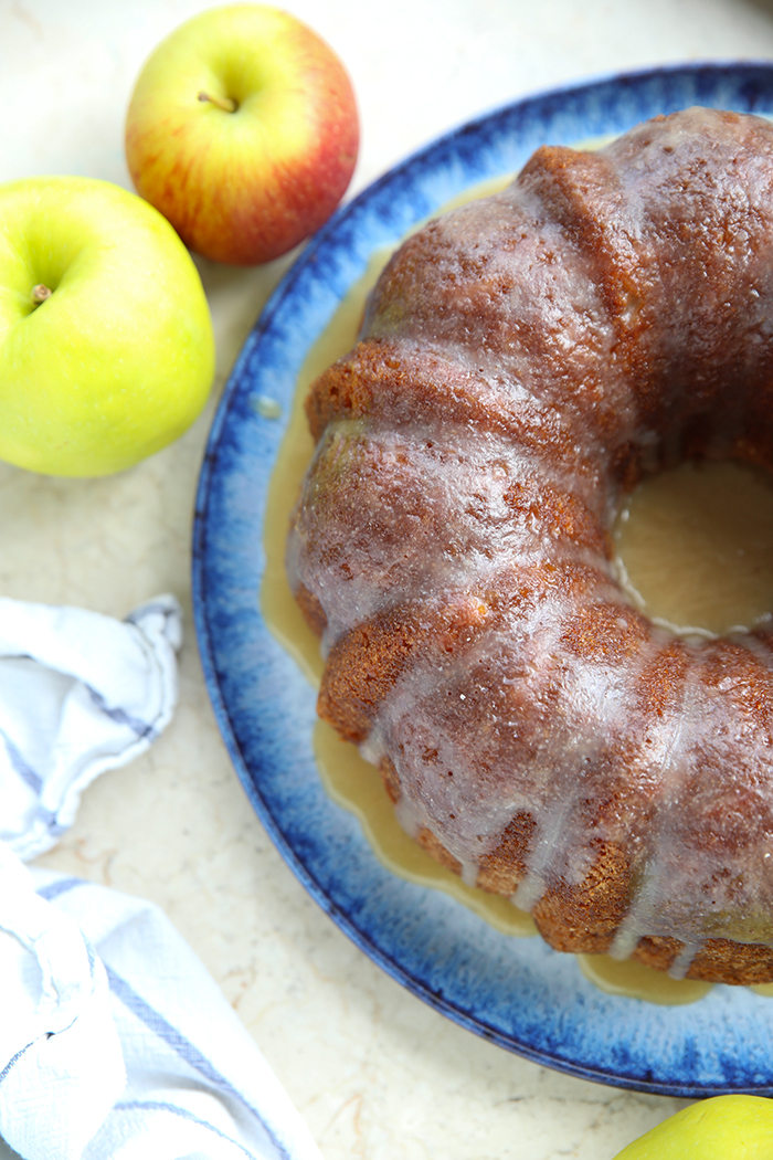 A partial picture of an apple bundt pound cake with two apples in the upper left corner and a dish towel in the bottom left corner.