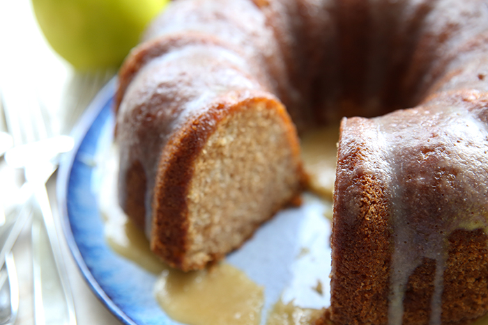 A slice cut out of an apple pound cake with caramel glaze on a round plate. Faded on the left of the plate are a couple of forks and above an apple.