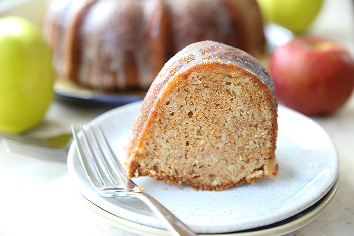 A serving of apple cake on two plates with a fork on top of the plates. Faded in the background are a few apples around the apple bundt cake.