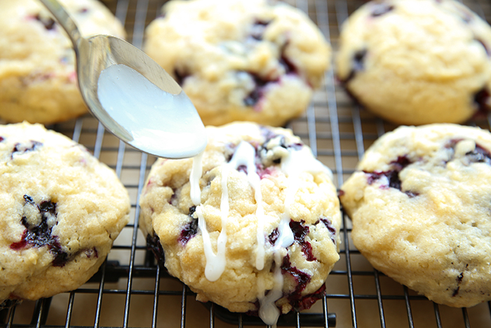 A wire cooling rack of blueberry muffin cookies with a spoon held above one cookie drizzling glaze on it.