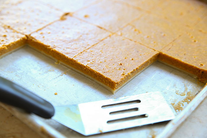 A half sheet pan of pumpkin slab pie cut into squares with several pieces missing and a serving spatula lying in the pan where the pieces are missing.