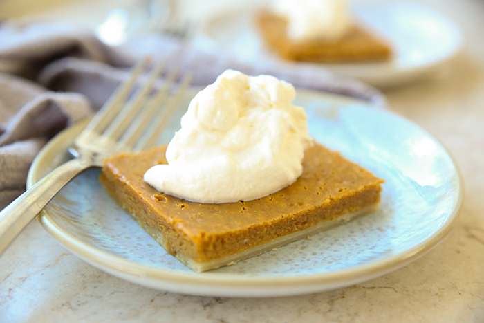 A slice of pumpkin slab pie with a dollop of whipping cream on a dessert plate with a fork on top of the plate. Faded in the background is a cloth tea towel and another piece of pie with whipping cream.