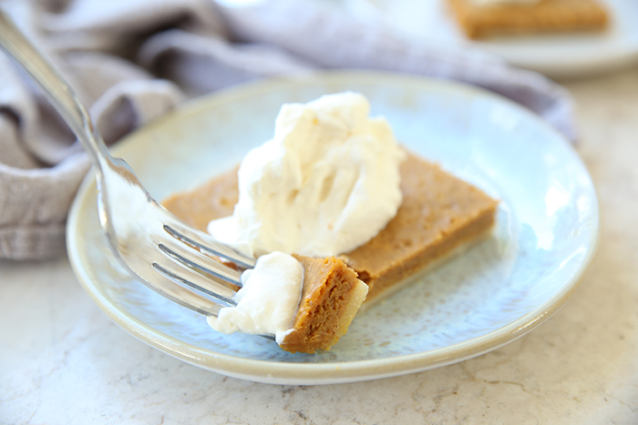 A square piece of sheet pan pumpkin pie topped with whipping cream on a small plate. A fork is held diagonally on the plate with a corner of the pie on it.