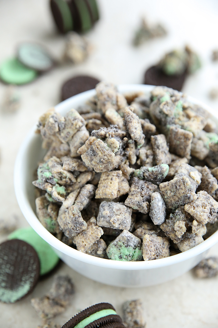 A cereal bowl full of Mint Oreo Muddy Buddies. Mint Oreos and some muddy buddies are scattered around the bowl.