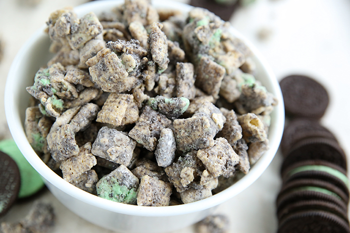 A small white bowl of Mint Oreo Muddy Buddies with a vertical stack of Mint Oreos lying on the right of the bowl.