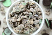 Mint Oreo cookies and mint muddy buddies scattered around a small white bowl of Mint Oreo Muddy Buddies.