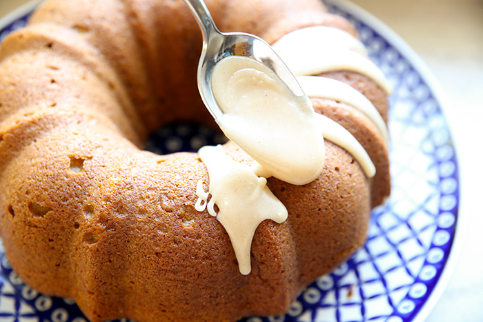 Pumpkin spice bundt cake on a round large Polish potter platter with a spoon held above placing brown butter icing on it.