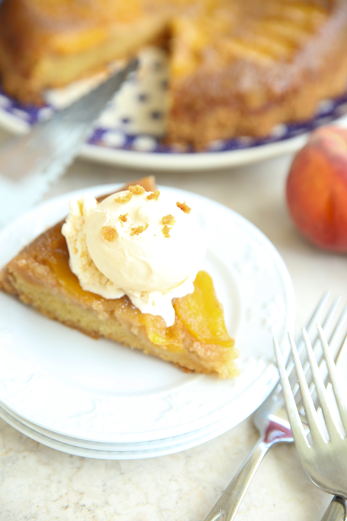 A slice of upside down peach cake topped with whipped cream sitting on three stacked small plates. In the bottom right corner are a couple of forks. Faded in the upper background is the upside down peach cake with a piece cut out and a peach and serving spatula.
