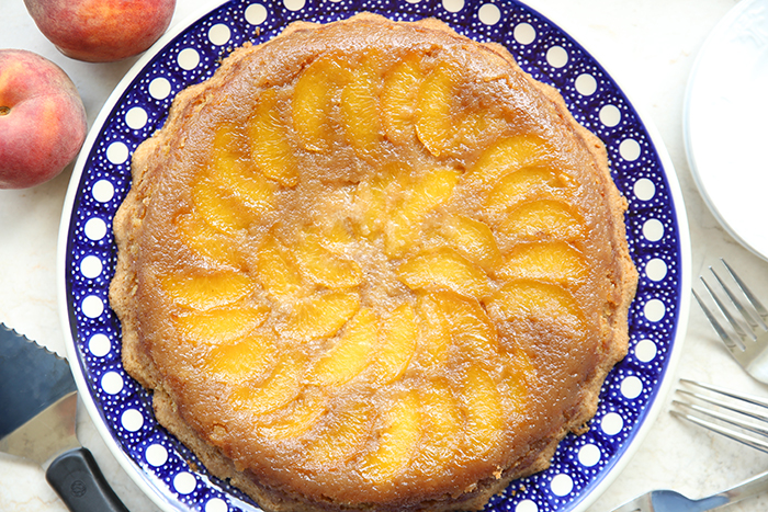 Upside Down Peach Cake on a round dark blue white polka-dotted Polish pottery dish. In the right corner are a couple of fork heads, and in the left corner a serving utensil. In the upper right corner are two whole peaches.