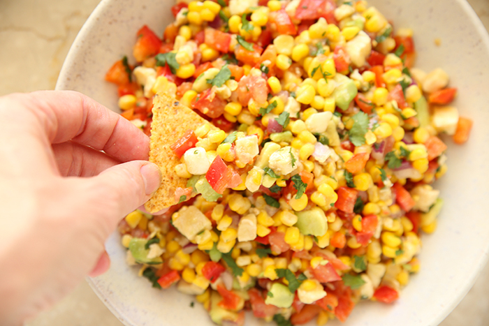 A hand held above a bowl of Janeth's Corn Salad Dip holding a chip with dip on it.