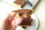 A chocolate peanut butter bar with a bite taken out of a corner held above a small plate with a couple of peanut butter bars and the corner of a sheet pan with a peanut butter bar and the handle of a serving spatula.