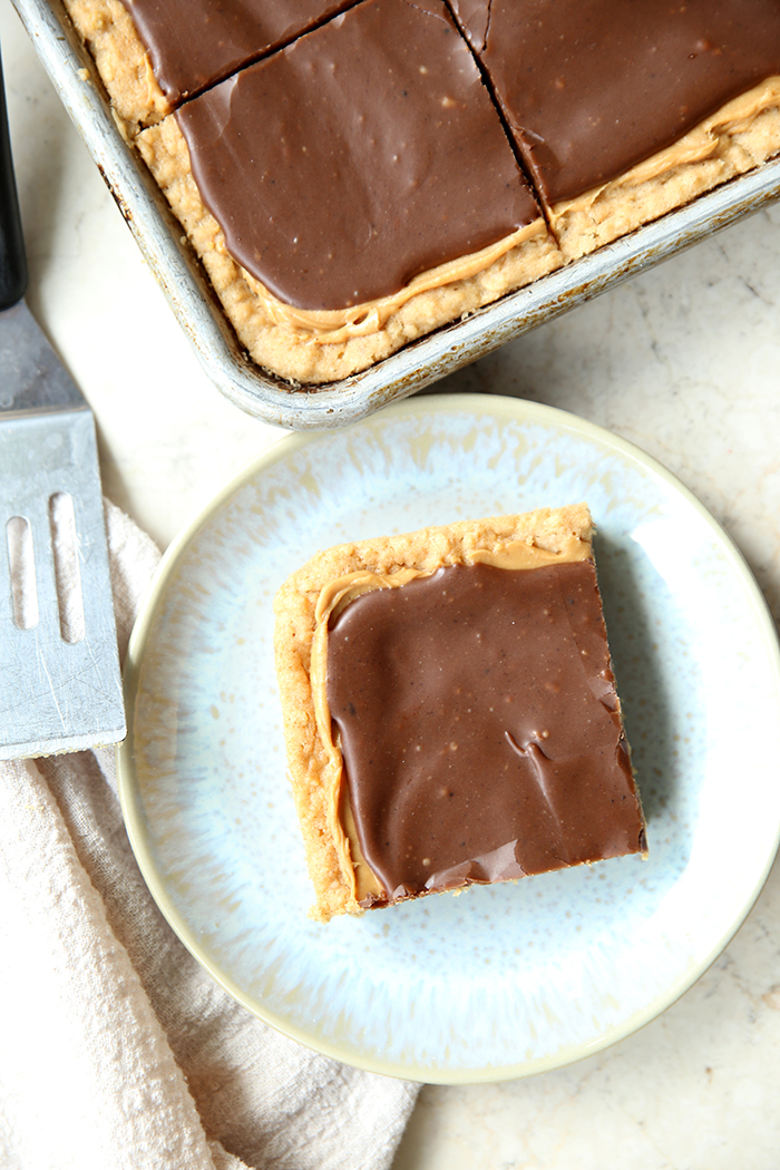 A square piece of lunch lady peanut butter bar on a small plate. Next to the plate is a serving spatula head and a cloth kitchen towel. Above the plate is the corner of a 18x13 sheet pan of cut peanut butter bars.