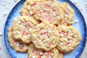 A large blue plate with a dark blue border filled with white chocolate peppermint cookies with white chocolate chips and peppermint pieces surrounding the plate.