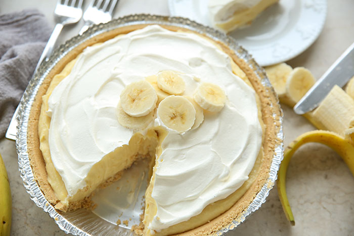 No-bake banana cream pie with a slice taken out. On the right is a peeled banana partially sliced with a knife. In the upper left corner are two forks next to a grey kitchen towel.