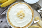 Easy no-bake banana cream pie with a grey kitchen towel to the left, a banana on the upper left, and a peeled and partial slice banana and a few forks on the right.