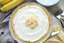 Easy no-bake banana cream pie with a grey kitchen towel to the left, a banana on the upper left, and a peeled and partial slice banana and a few forks on the right.
