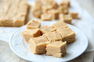 8 pieces of peanut butter fudge on a small plate. Faded in the background are more pieces of fudge and the leftover slab of fudge.