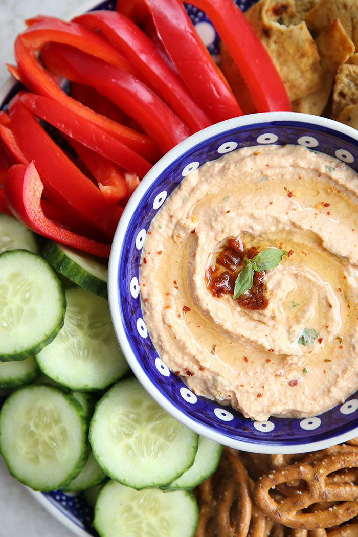 A bowl of sun dried tomato hummus sitting on a round plate surround by pretzels, pita chips, sliced red bell pepper and sliced cucumbers.