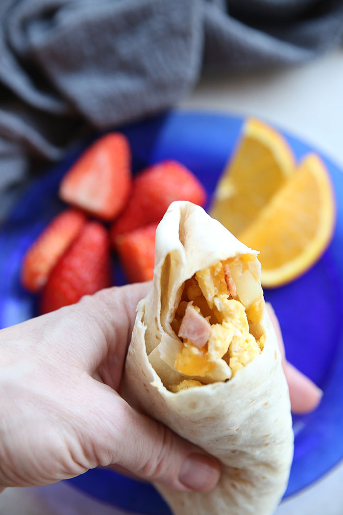 A ham, potato and egg breakfast burrito with a bite taken out of it held above a plate with sliced strawberries and orange slices and a kitchen towel.