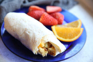 An egg, ham and potato burrito with a bite taken out on a blue dinner plate with sliced strawberries and orange slices.