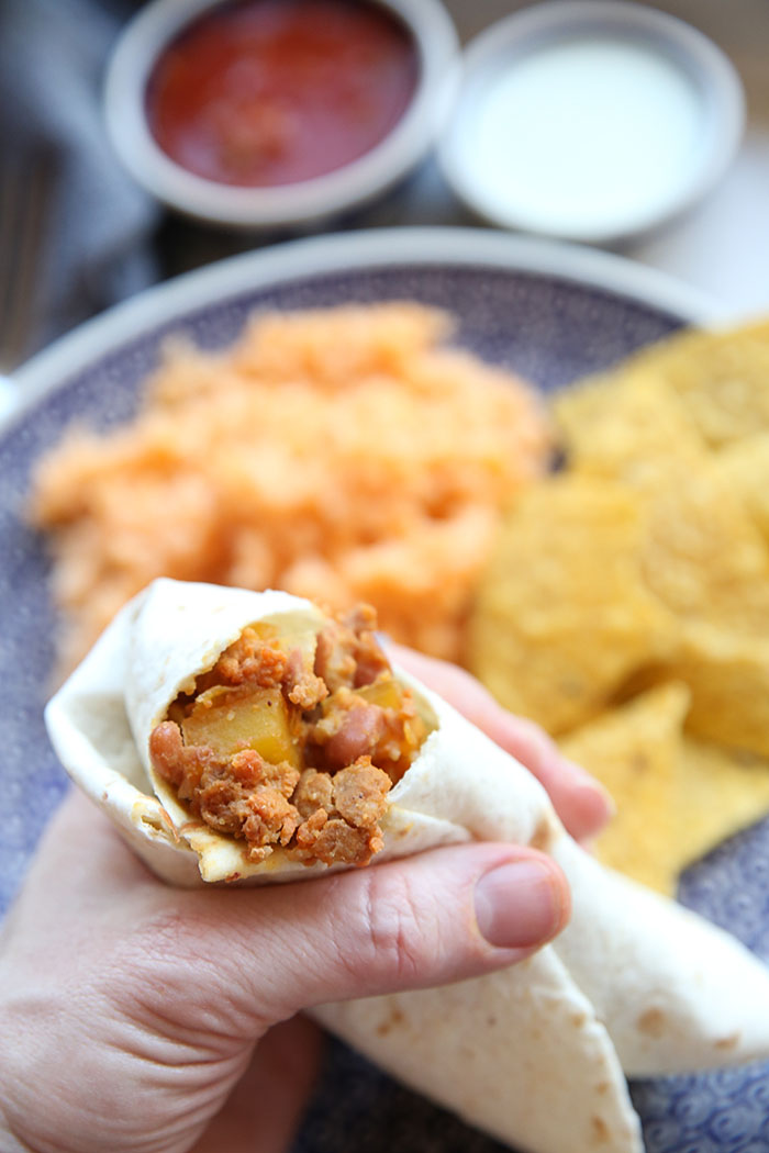 Chorizo, potato, and pinto bean burrito with a bite taken out held above a plate with Spanish rice and corn chips, and a small bowl of salsa and a small bowl of sour cream.