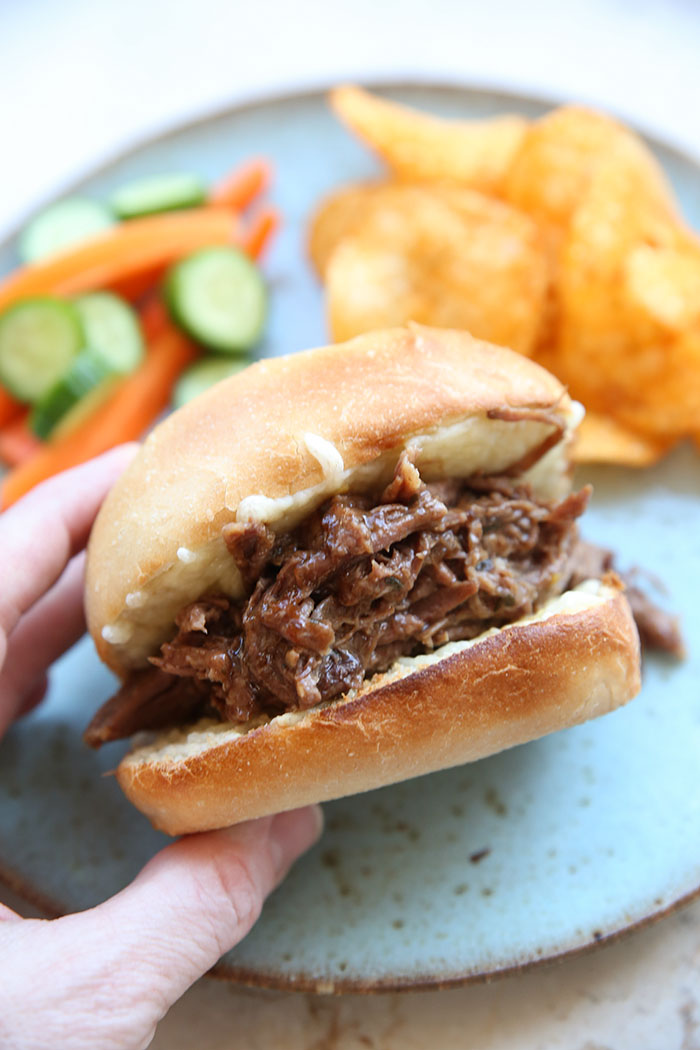 Shredded Mississippi pot roast in a bun with melted cheese held diagonally on a plate with barbecue chips, sliced carrots and sliced cucumbers.