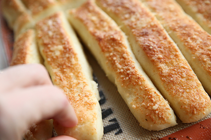 Parmesan garlic breadsticks on a cookie sheet with a silpat mat with a hand pulling a breadstick away.