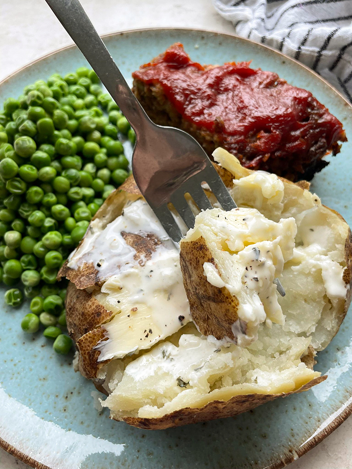 A dinner plate with a serving of meatloaf and green peas next to a baked Russet potato cut open with butter, salt and pepper with a fork held vertically with a piece of baked potato on it.