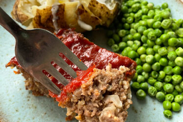 A rectangle slice of meatloaf with a fork angled from the left inserted into a corner of the meatloaf sitting on a plate with a cut open baked potato and a serving of green peas.